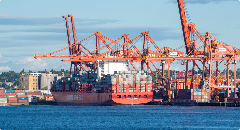 A large cargo ship with shipping containers is docked at a British Columbia port, featuring orange cranes and stacked containers, highlighting trade infrastructure.