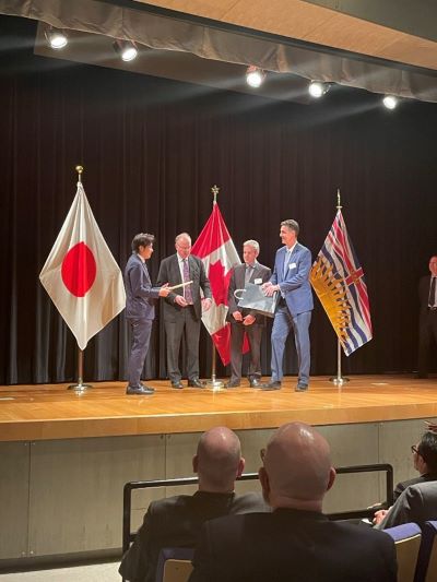 ステージの上で日本、カナダ、BCの国・州旗の横で握手している4人。