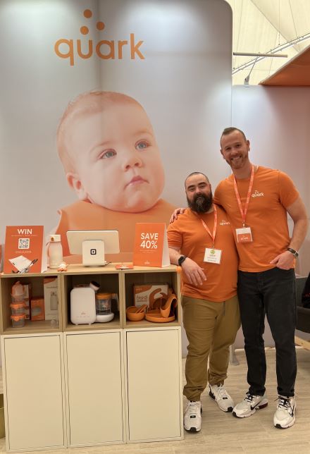 展示会ブースで商品の横に立つQuarkの共同創業者、セネズとグリンスカス。ロゴの背面に赤ちゃんの写真。